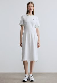 Λευκό βαμβακερό φόρεμα με κοντά μανίκια, πράσινες λεπτομέρειες στον γιακά και τα μανίκια, με χαλαρή εφαρμογή. Φορεμένο με λευκά και μαύρα αθλητικά παπούτσια.