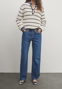 Pull en maille crème à rayures foncées, superposé à une chemise à carreaux bleue, associé à un jean denim bleu à jambes larges et des ballerines argentées.