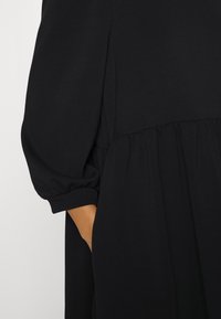 Black dress with a round neckline, long puff sleeves, and a gathered waistline. Soft texture, knee-length, with side pockets.