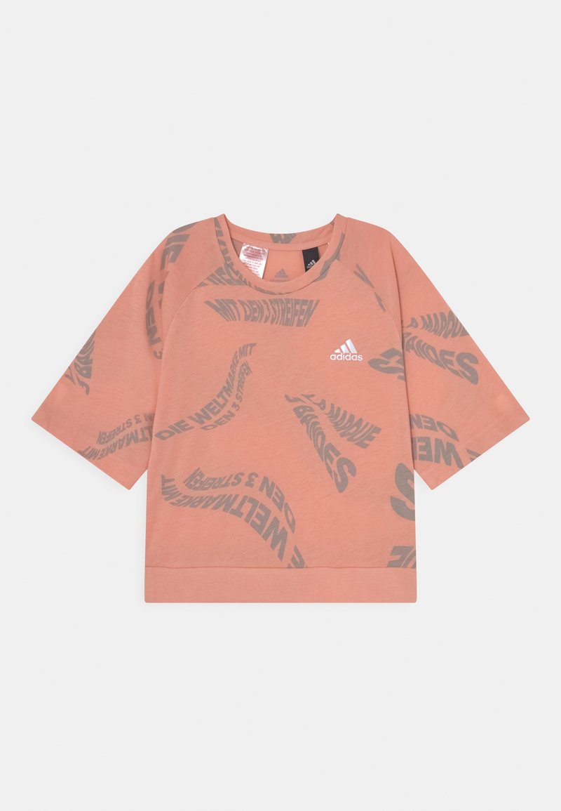 Koszulka z krótkim rękawem i okrągłym dekoltem w jasnym brzoskwiniowym kolorze, z szarymi, zakrzywionymi wzorami tekstowymi oraz logo adidas na przodzie. Miękka, lekka tkanina.