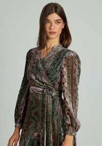 Wickeldress mit einem Muster aus Grün, Lila und Gold. Lange Ärmel, geraffte Taille mit passendem Gürtel, strukturierten Samtstoff.