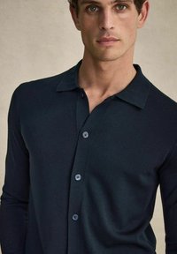 Mörk marinblå långärmad skjorta med krage, tillverkad av ett slätt tyg. Har en knappslå med kontrasterande blå knappar.