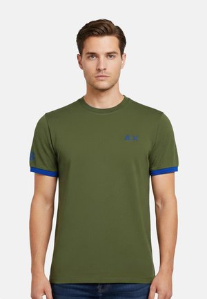 Uomo che indossa una t-shirt verde oliva a maniche corte con rifinitura blu sulle maniche e un piccolo logo blu sul petto, in piedi contro uno sfondo semplice.