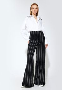 Camisa blanca de manga larga con cuello en contraste, combinada con pantalones negros de pierna ancha con rayas verticales blancas. Textura de tela suave.