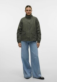 Olijfgroen jack met hoge kraag, drukknoopsluiting en elastische boorden. Voorzien van zijzakken en een trekkoord aan de onderrand. Graag gedragen met blauwe wijde jeans.
