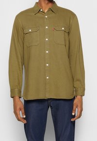Oliwkowa koszula z długim rękawem wykonana z bawełny, z dwoma przednimi kieszeniami, zapięciem na guziki i zaokrąglonym dołem. Białe guziki i szczegół z czerwonym logo.