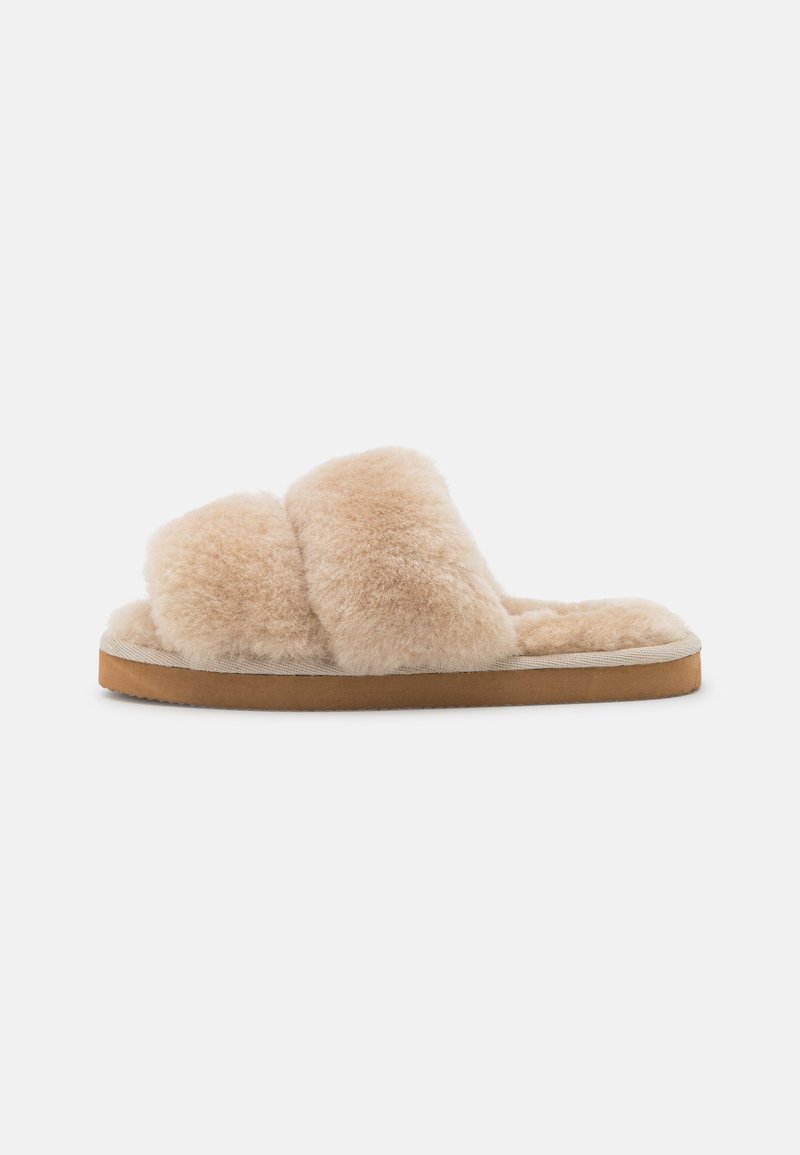 Flauschige beige Schlüpfschuhe mit zwei breiten Kreuzriemen, einem weichen Innenfutter und einer braunen Gummisohle. Minimalistisches Design, ohne sichtbare Beschläge.