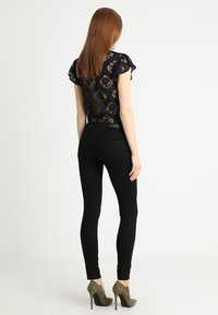 Top de encaje negro con mangas cortas y volantes, combinado con jeans ajustados negros de cintura alta. La modelo lleva zapatos de tacón altos en color verde oliva.