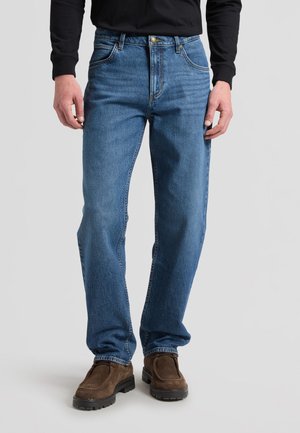 Homme portant un jean bleu coupe droite avec des chaussures en daim marron et une chemise noire à manches longues, debout devant un fond clair uni.