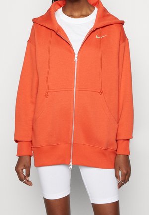 Kobieta ubrana w jaskrawą pomarańczową bluzę z kapturem na zamek z małym metalicznym logo Nike, pod spodem biała koszulka i białe szorty rowerowe.