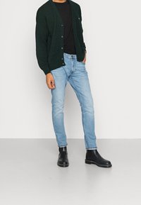 Mann trägt eine dunkelgrüne Strickjacke über einem schwarzen Shirt, hellblaue Slim-Fit-Jeans und schwarze Lederstiefeletten, steht auf einem grauen Boden.