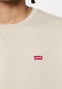 Felpa beige a girocollo con una piccola patch rossa del logo Levi's sul lato sinistro del petto. Tessuto morbido con una sottile texture e colletto a coste.