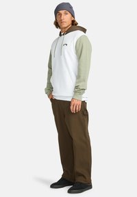 Hoodie met een witte lichaam, olijfgroene mouwen en bruine accenten; gecombineerd met een paar bruine broeken en zwarte schoenen. Gestructureerde gebreide beanie.