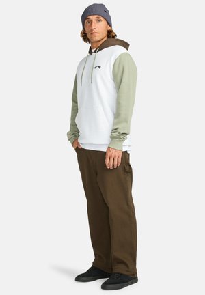 Hoodie met een witte lichaam, olijfgroene mouwen en bruine accenten; gecombineerd met een paar bruine broeken en zwarte schoenen. Gestructureerde gebreide beanie.
