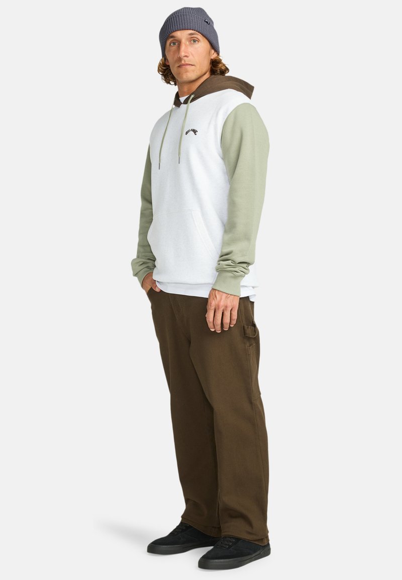 Hoodie met een witte lichaam, olijfgroene mouwen en bruine accenten; gecombineerd met een paar bruine broeken en zwarte schoenen. Gestructureerde gebreide beanie.