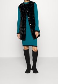 Teal-randig klänning med en svart faux fur-väst med stora knappar. Modellen bär knähöga svarta tygstövlar med en kraftig klack.