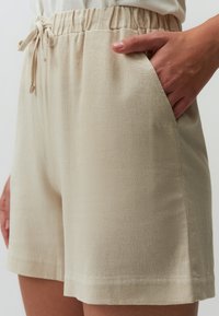 Beige shorts met een elastische tailleband, trekkoord en zijzakken. Zachte textuur en eenvoudig ontwerp, afgewerkt op kniehoogte.