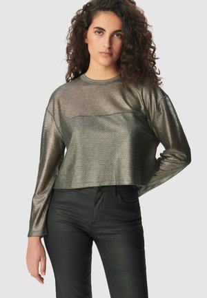 Donna con capelli ricci che indossa un top a maniche lunghe in rete metallica e pantaloni neri aderenti, in piedi con una mano sulla vita su uno sfondo neutro.