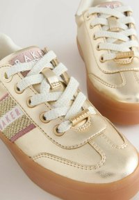 Kuldse nahast kingad valgete paeltega, roosa ja kuldse kudumisega külgtriibud ning paksud beežid kummitaldad kerge tausta peal.