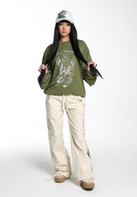 Olive szara, oversize bluza z białym nadrukiem graficznym, w połączeniu z kremowymi spodniami cargo z bocznymi rozcięciami i haftami w kwiatowy wzór. Biała czapka.