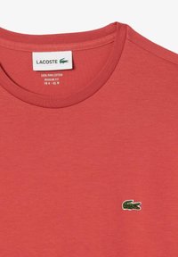 Röd Pima-bomull t-shirt med rund halsringning och en liten grön krokodil-logotyp broderad på vänster bröstområde. Normal passform.