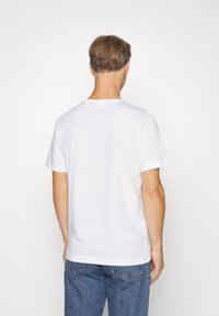 Férfi háttal, egyszerű fehér rövid ujjú pólót és kék farmert visel, fehér háttér előtt.