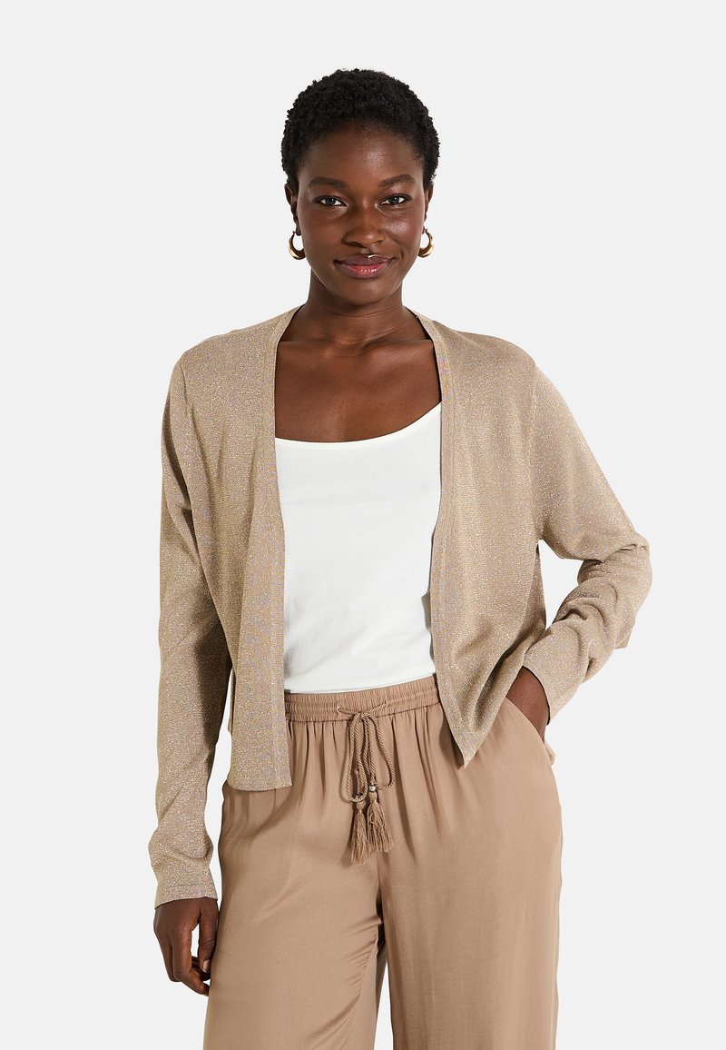 Cárdigan color beige claro con un sutil brillo, de frente abierto, mangas largas, combinado con una blusa blanca y pantalones beige con cordón.