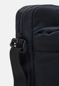 Marinblå tygväska med dragkedja, som har en strukturerad yta, justerbar svartrem och en cirkulär detalj i metall.