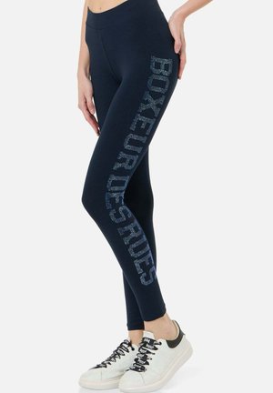 Frau trägt marineblaue Leggings mit "BOXEUR DES RUES" aus Strasssteinen an der Seite, kombiniert mit weißen Sneakers.