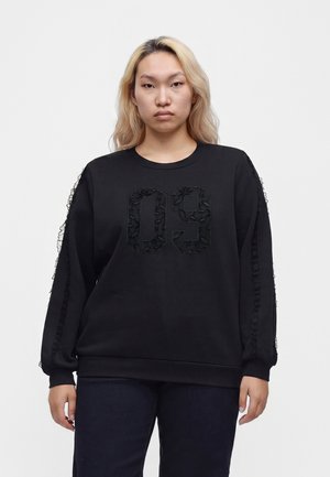 Person med langt blondt hår iført en sort sweatshirt med tekstureret "09" foran og dekorative striber ned ad begge ærmer.