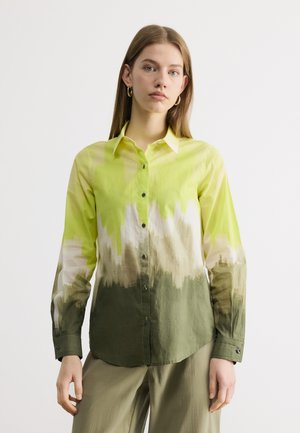 Νεαρή γυναίκα που φοράει μακρυμάνικο πουκάμισο με κουμπιά, με μοτίβα σε σχήμα κλίσης κίτρινου, λευκού και πράσινου, συνδυασμένο με ανοιχτό πράσινο παντελόνι.