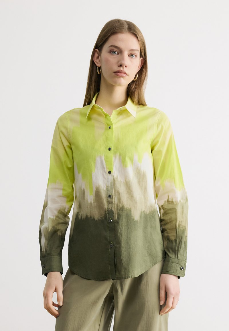 Fiatal nő hosszú ujjú, gombos ingben, amely sárga, fehér és zöld átmenetes mintákkal díszített, világos olívazöld nadrággal kombinálva.