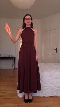 Γυναίκα με αμάνικο μπαordo φόρεμα και γυαλιά, όρθια ξυπόλυτη σε ξύλινο πάτωμα, σηκώνοντας το αριστερό χέρι σε χαιρετισμό.