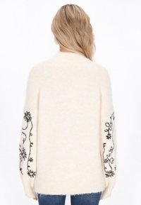 Kremowy, oversize sweter z czarną haftowaną florą na rękawach, z prążkowanym kołnierzem i mankietami, wykonany z miękkiej, teksturowanej dzianiny.