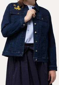 Chaqueta de mezclilla azul oscuro con cierres de botón, dos bolsillos en el pecho y bordado floral en el hombro. Llevada sobre una camisa a rayas.