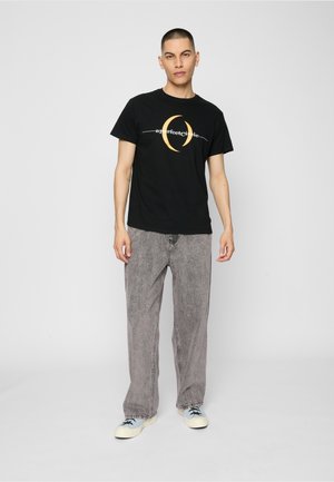 Homme debout portant un t-shirt noir à motif avec le texte « a perfect circle », un jean gris ample et des baskets blanches avec des lacets noirs.