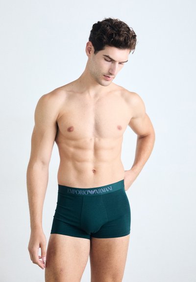 Temnozeleni bombažni bokserji sivoj pasu Emporio Armani, ki se odlikujejo po tesnem prileganju in gladki teksturi, oblečeni na moškem modelu.