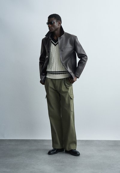 Άντρας που φοράει μαύρα γυαλιά ηλίου, σκούρο δερμάτινο μπουφάν, μπεζ πουλόβερ με πλεκτό σχέδιο και μαύρες ρίγες, χακί παντελόνι cargo και μαύρα παπούτσια, στέκεται σε εσωτερικό χώρο.