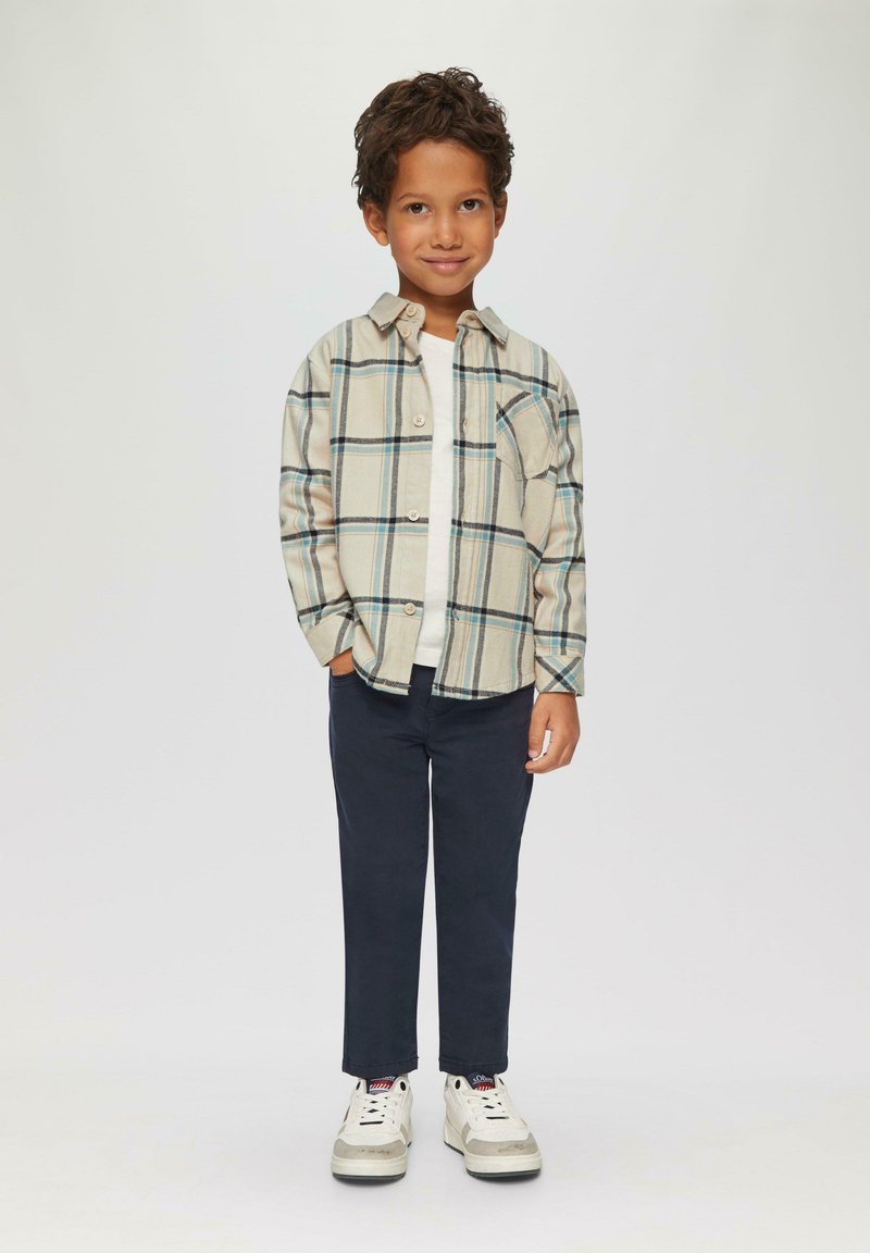 Lichtbeige geruite shirt met groene en blauwe lijnen, gecombineerd met donkerblauwe broek en witte sneakers. Korte krullende haren. Casual look.