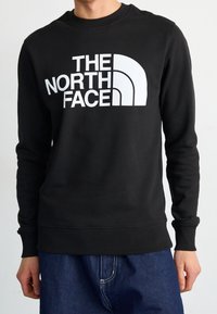 Svart sweatshirt med en stor vit tryckt logotyp som säger "THE NORTH FACE," med ribbstickade mudd och nederkant. Slät textur.