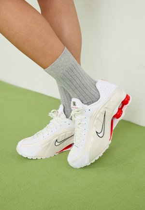 Benen met grijze geribde sokken en witte Nike-sneakers met rode veerachtige zolelementen op een groene vloer bij een witte muur.