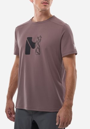Homme portant une chemise mauve à manches courtes avec un logo abstrait noir et à motifs sur la poitrine et un pantalon gris.