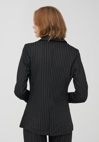 Blazer nero con righe sottili e vestibilità sartoriale, dotato di revers a punta, maniche lunghe e spacco posteriore. Texture liscia e design strutturato.