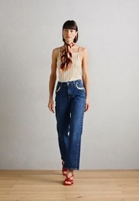 Top plissettato beige chiaro, jeans blu a vita alta con accenti argentati e sandali rossi con cinturini. La modella si trova su un pavimento di legno contro un muro grigio.