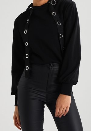 Personne portant un haut noir à manches longues avec des sangles à œillets argentés et un pantalon noir en cuir taille haute, debout devant un fond uni.