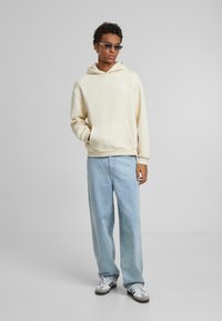 Cremefarvet hættetrøje med frontlomme, hvid tekst og snoretræk i hætten. Lys blå bukser med vide ben og sort-hvide sneakers fuldender outfittet.