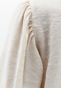 Beige, gestructureerde stof met zachte plooien langs de mouw die zachtjes hangen. Het materiaal lijkt lichtgewicht en iets gekreukeld.