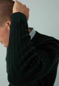 Mann mit erhobener Hand nahe dem Kopf, der einen dunkelgrünen strukturierten Strickpullover und ein hellblaues Hemd mit Kragen trägt.