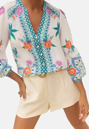 Woman wearing long-sleeve floral blouse with teal geometric trim and cream high-waisted shorts with a ring belt, posing with hands resting on hips.