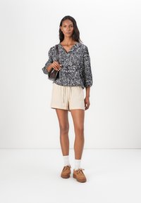 Svart og hvit mønstrete bluse med 3/4 ermer, parret med beige shorts. Brune platå-sandaler og liten mørk skulderveske.